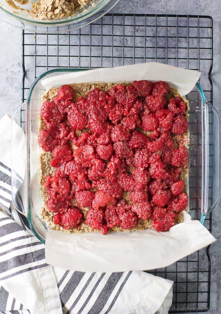 Almond Oat Raspberry Bar Recipe 一个羊皮纸衬里的平底锅,里面有杏仁覆盆子燕麦条,一些羊皮纸悬在外面