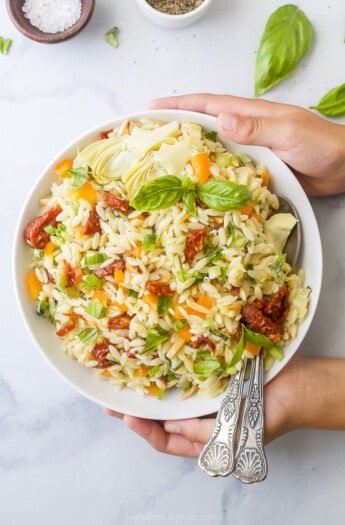 Two Hands Holding a Bowl of Mediterranean Salad with Two Spoons Inside